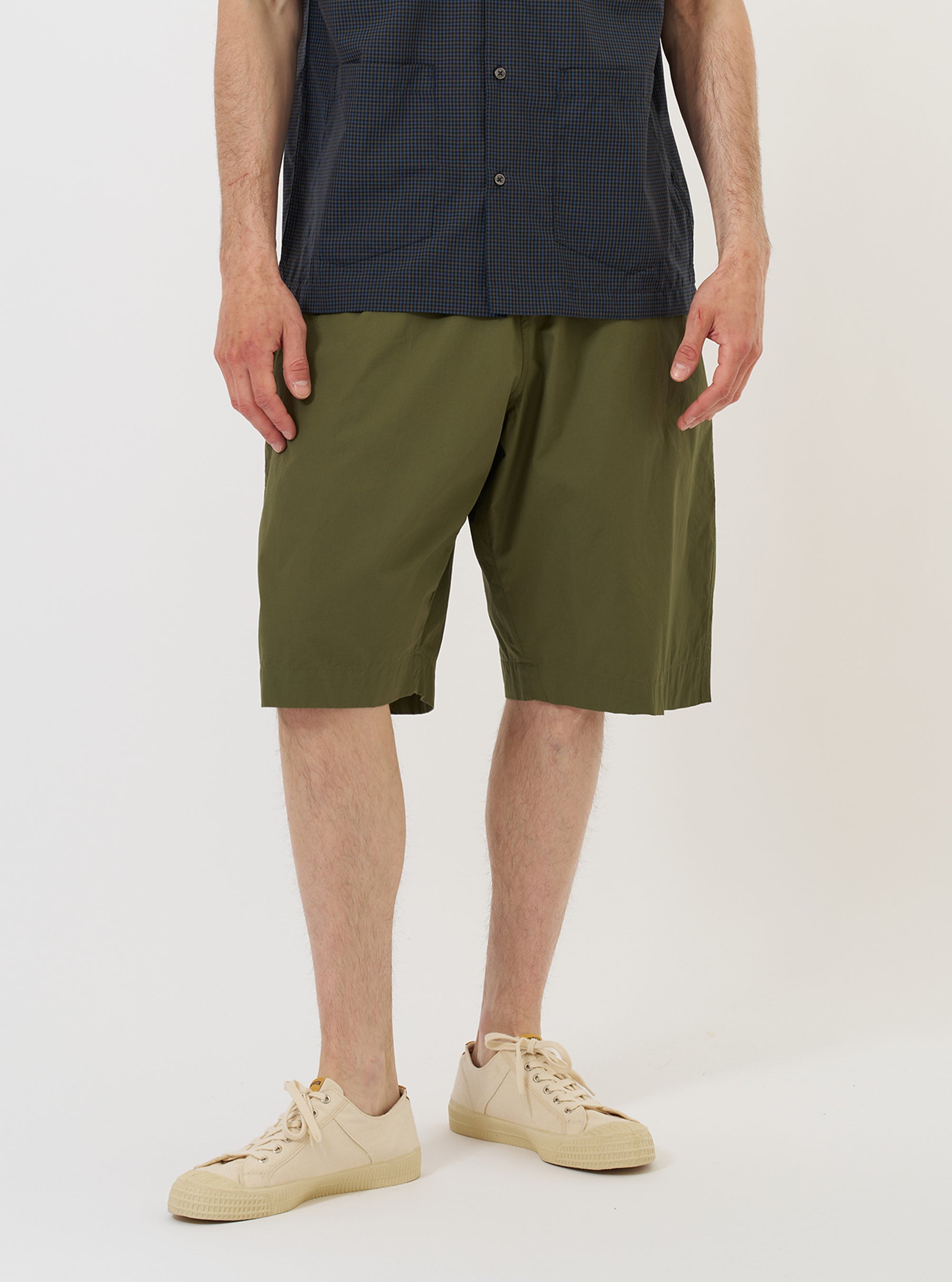 A model is wearing the Olive Broadcloth Vacation Short against a plain white background. The image shows the shorts from the waist down, highlighting the olive green cotton fabric, a relaxed fit, and a side slip pocket. The model is standing straight and the garment is paired with dark-colored shoes.