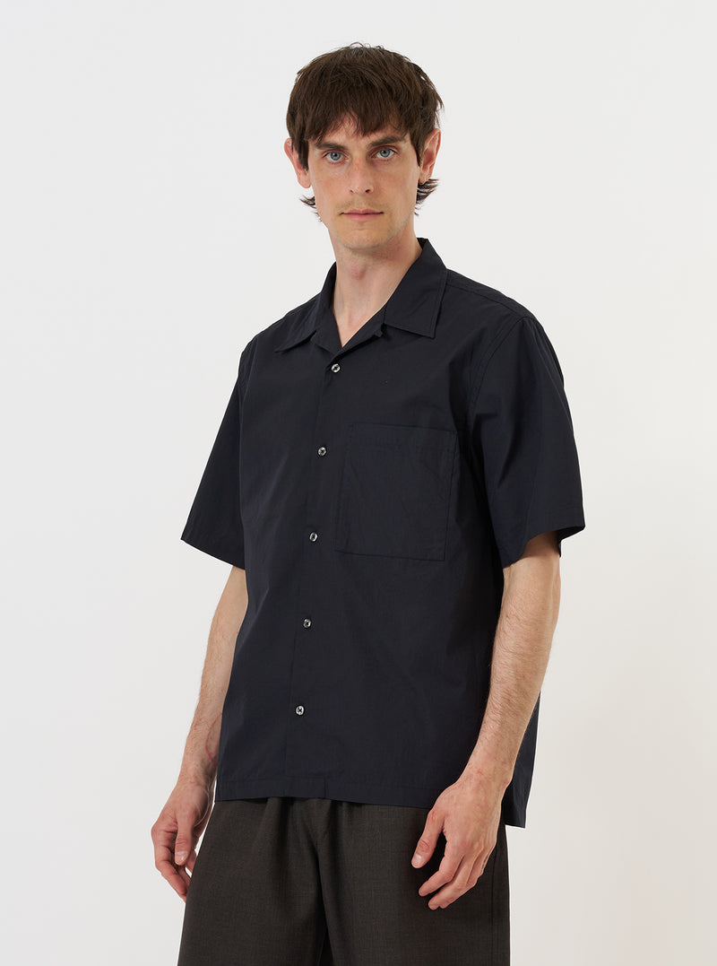 A model is wearing the Navy Broadcloth Camp Shirt against a plain white background. The short-sleeved navy blue shirt features a camp collar, a button-down front, and two large patch pockets at the waist. The model is standing and facing the camera, showcasing the shirt's relaxed fit and utilitarian design.