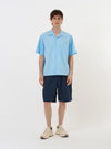 The Pale Blue Broadcloth Camp Shirt is shown laid flat against a solid white background. The shirt features a one-piece camp collar, a single large chest pocket on the left side, and a button-down front with a loop closure at the top. The garment has short sleeves and a straight hem with side splits.