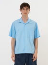 A model is wearing the Pale Blue Broadcloth Camp Shirt while standing against a plain white background. The short-sleeve, light blue shirt features a loose fit, a one-piece camp collar, and a large chest pocket on the left side. The garment is shown buttoned up and paired with dark-colored trousers.