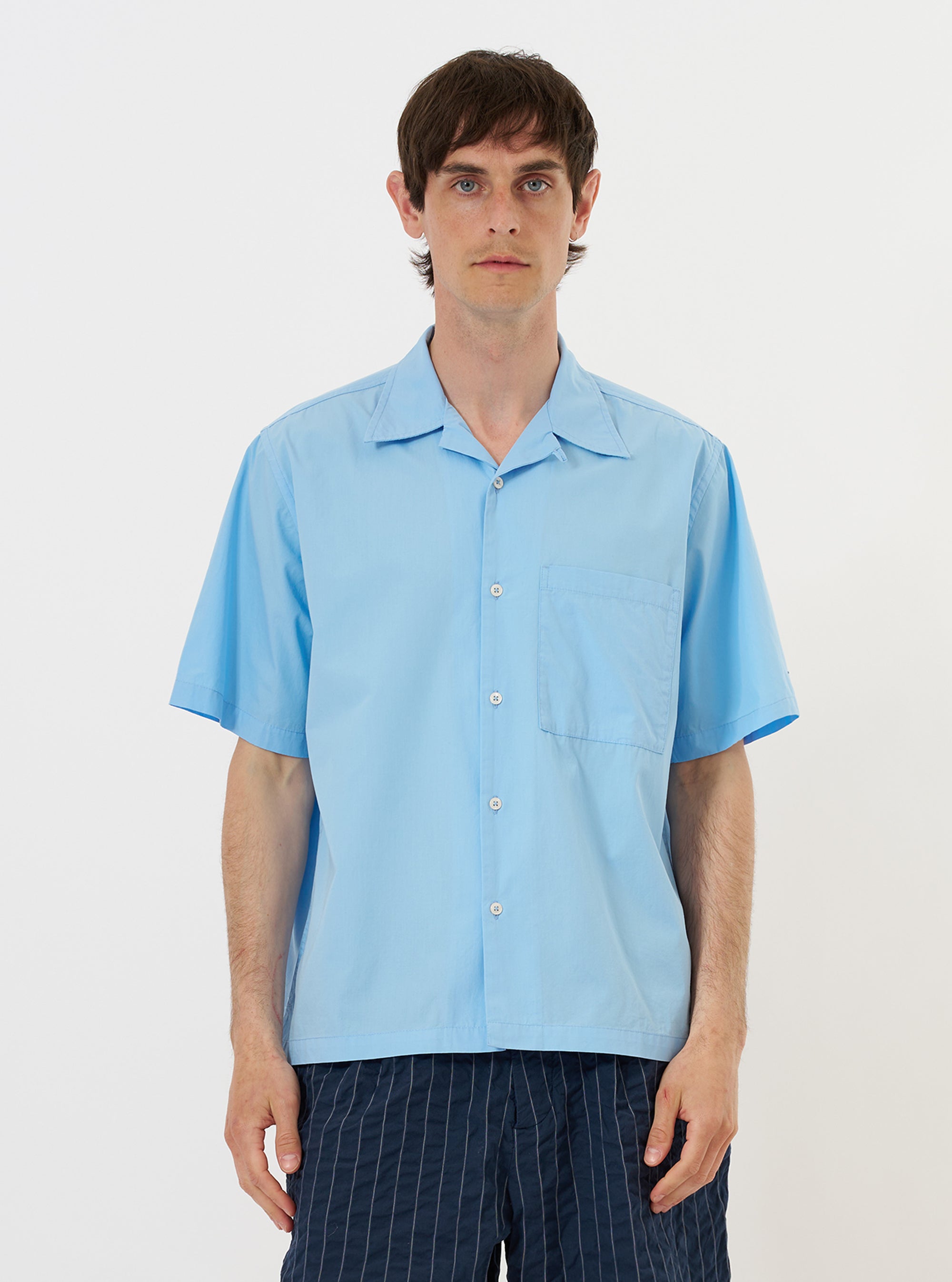 A model is wearing the Pale Blue Broadcloth Camp Shirt while standing against a plain white background. The short-sleeve, light blue shirt features a loose fit, a one-piece camp collar, and a large chest pocket on the left side. The garment is shown buttoned up and paired with dark-colored trousers.
