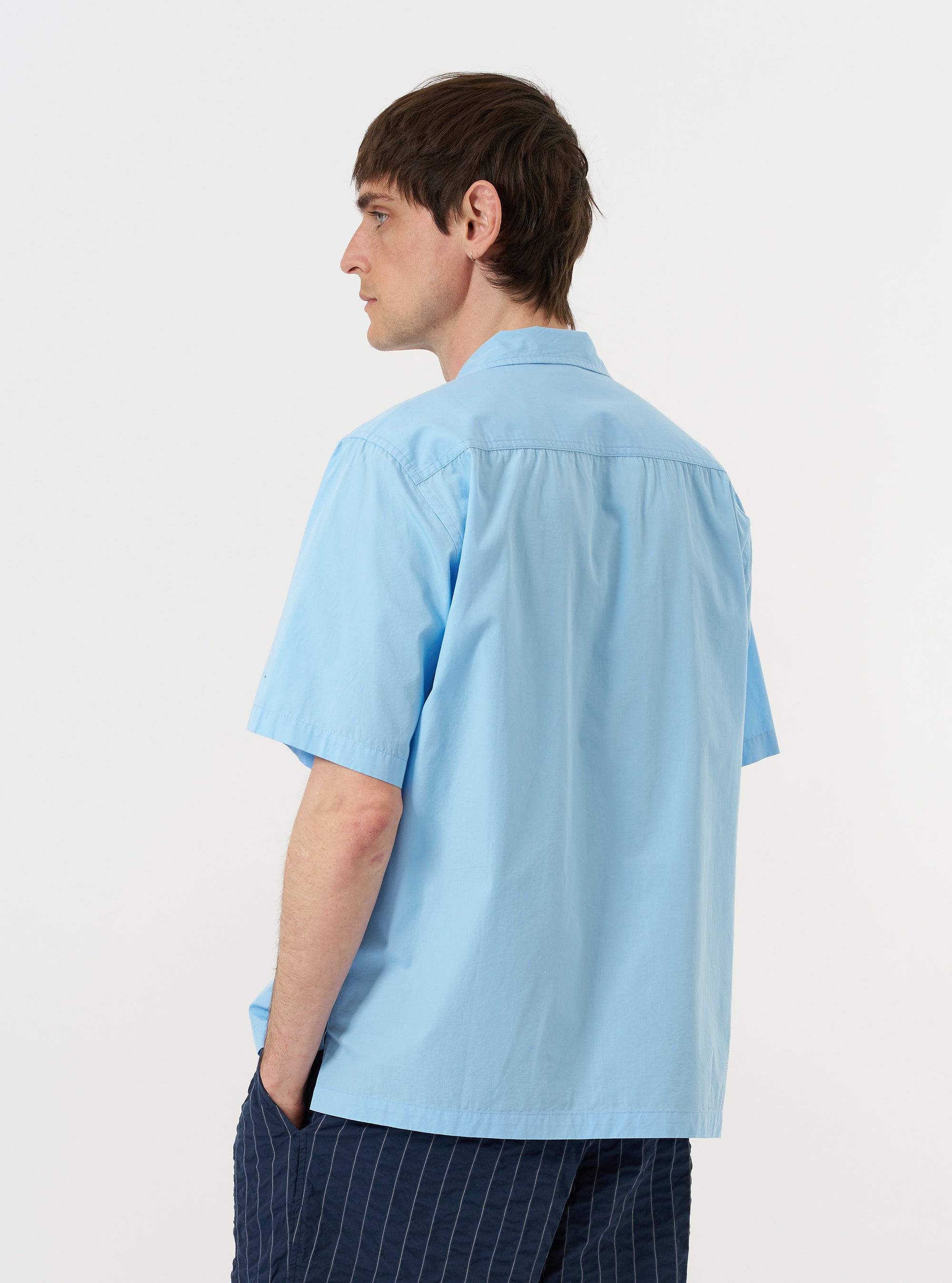 A model is wearing the Pale Blue Broadcloth Camp Shirt against a plain white background. The shirt is shown from the back, highlighting the back yoke with small pleat details and the straight-cut hem. The model has their hands by their sides, and the light blue, short-sleeve garment features a loose, relaxed fit.