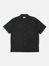 The Black Broadcloth Camp Shirt is shown laid flat against a solid white background. The shirt features a one-piece camp collar, a button-down front, and a single large chest pocket on the left side. The short-sleeve garment is made from a smooth, black broadcloth cotton fabric.