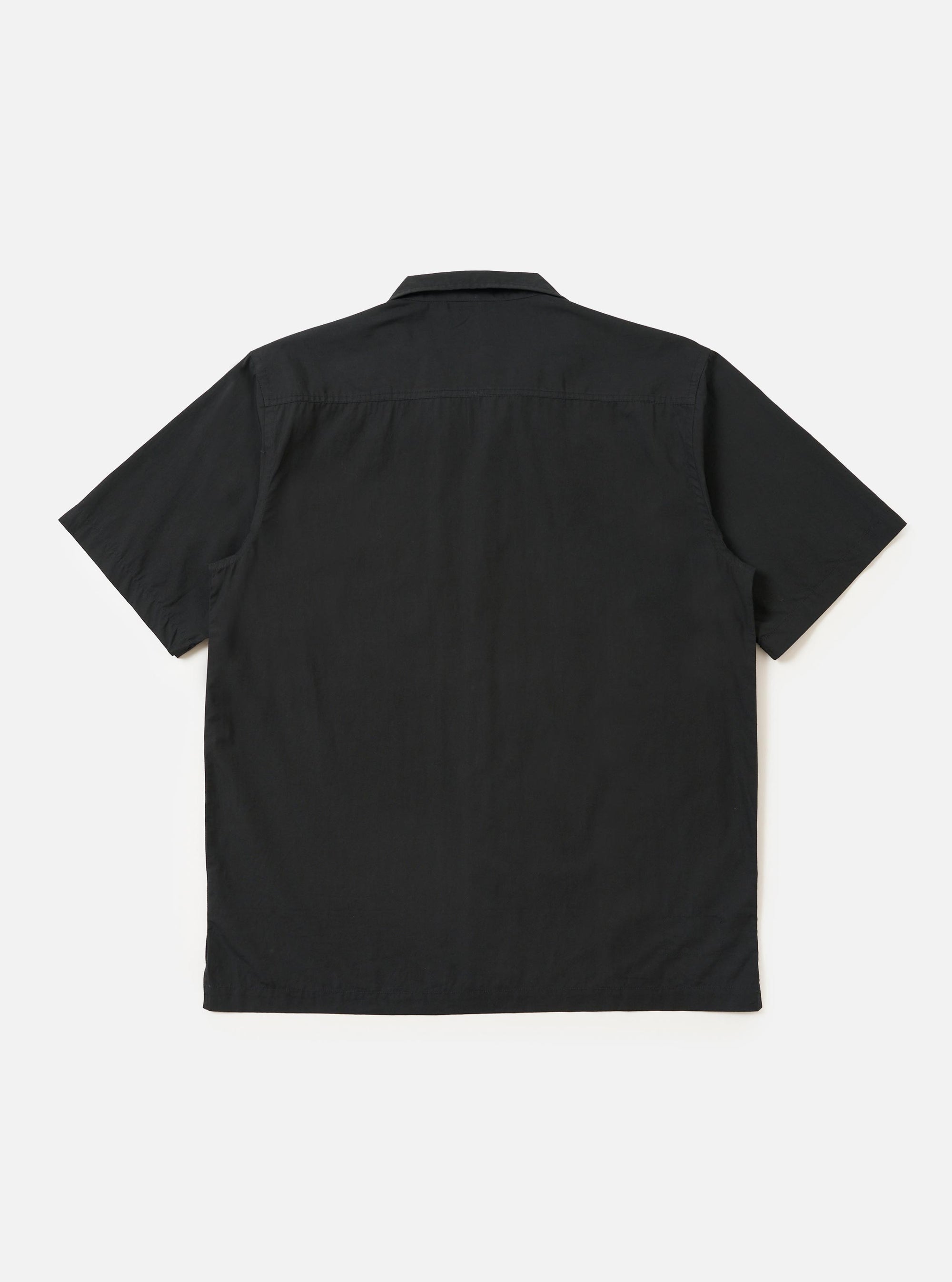 The Black Broadcloth Camp Shirt is shown from the back, laid flat against a plain white background. The image highlights the shirt's back yoke with small pleats and a straight, elongated hem with side splits.