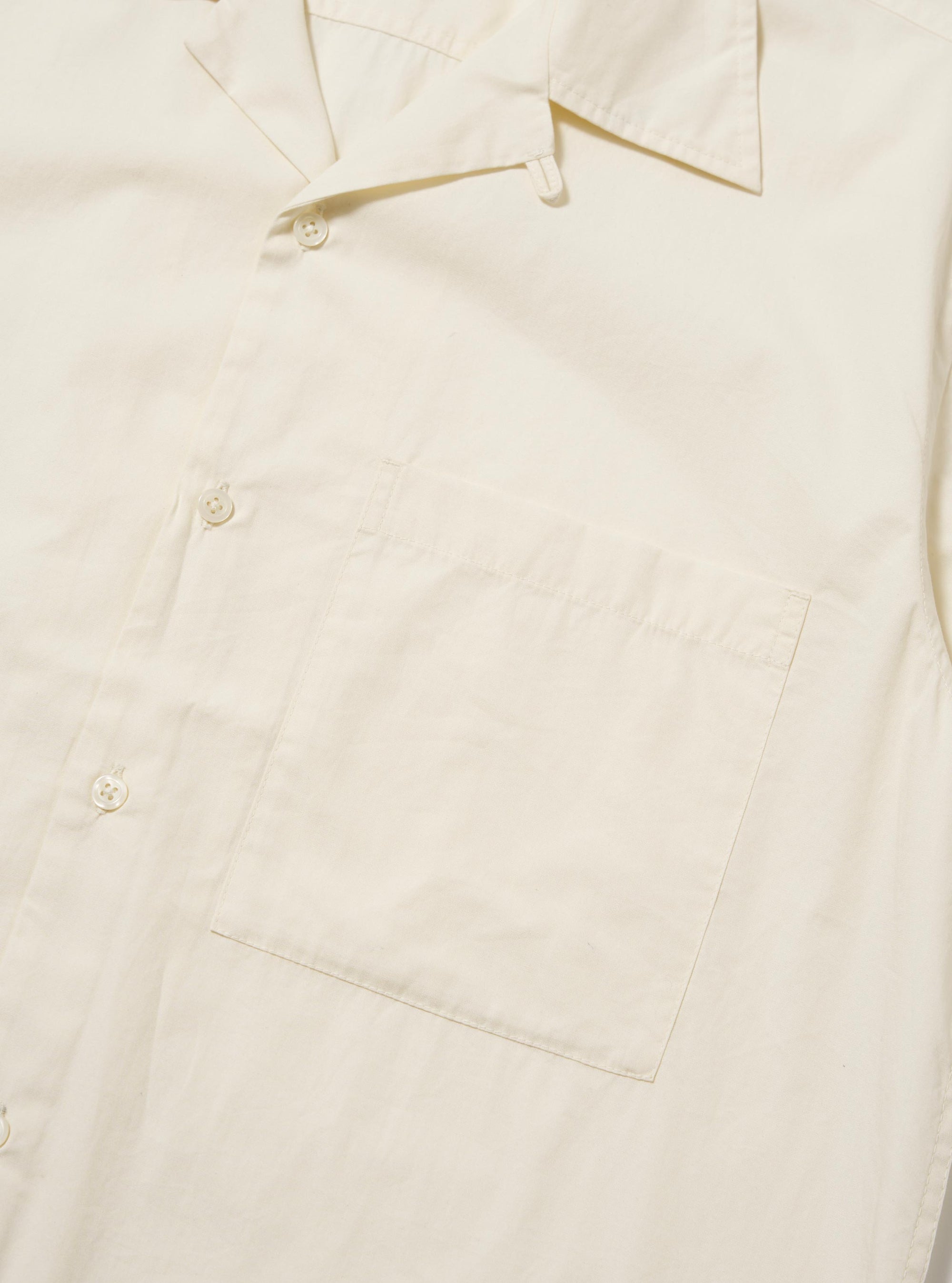 The Ecru Broadcloth Camp Shirt is shown from the back, laid flat against a plain white background. The image highlights the horizontal back yoke with two small pleats for added volume and the straight hem with side splits. The short sleeves are laid out to the sides, and