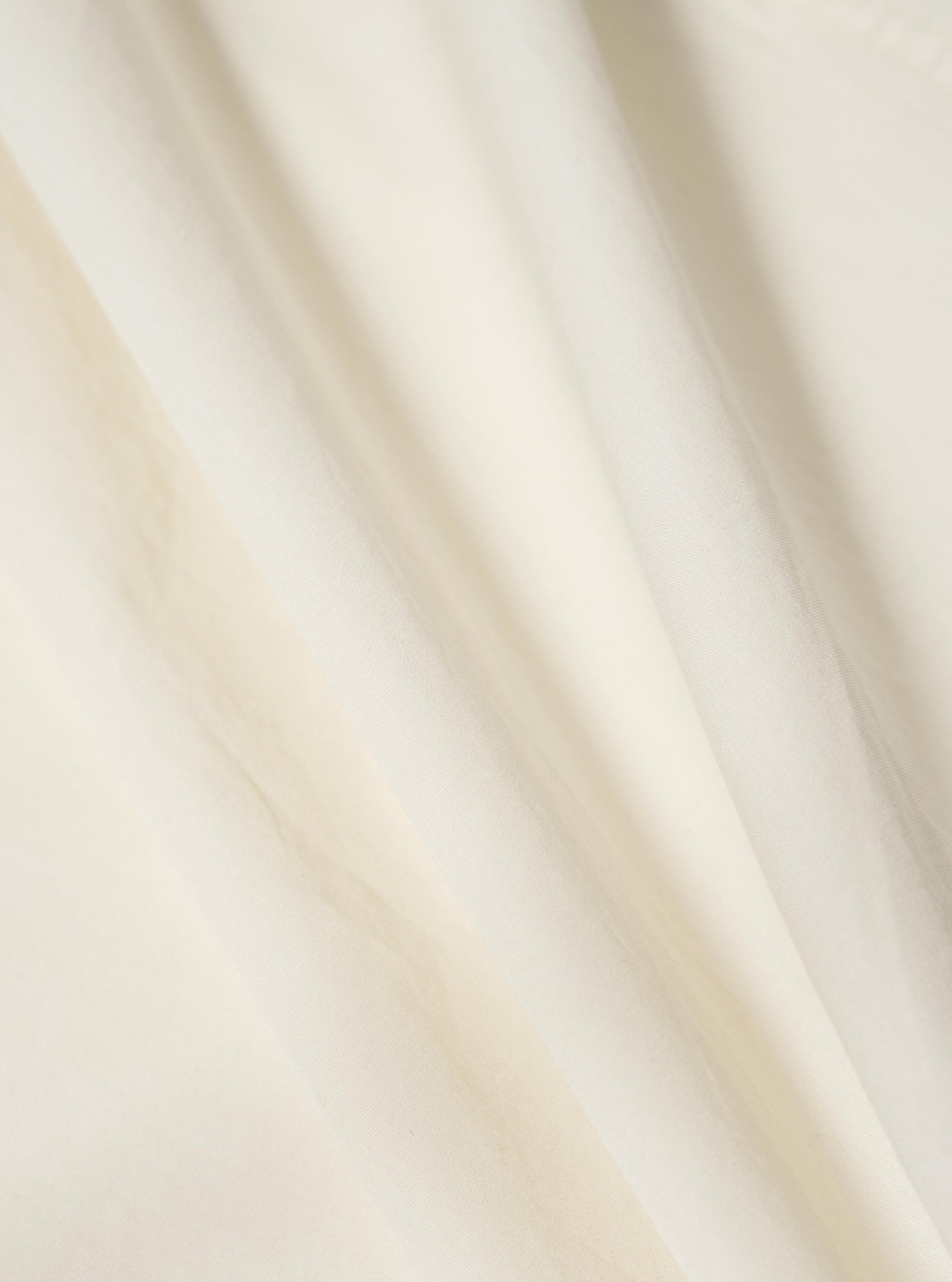 A close-up fabric swatch of the Ecru Broadcloth Camp Shirt displays the fine, smooth weave and off-white color of the broadcloth material. The image focuses on the texture and uniform finish of the cotton fabric.