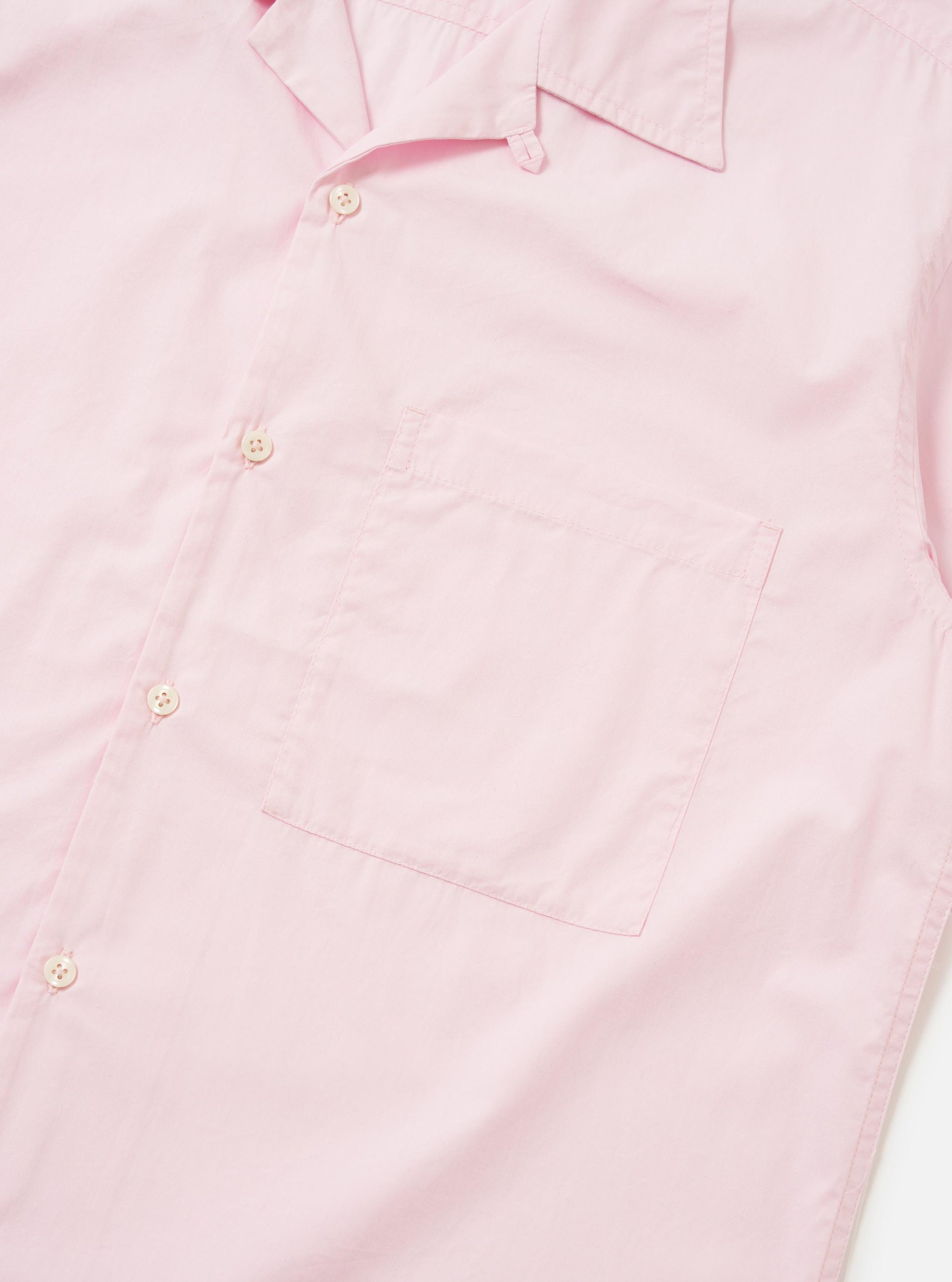 A Pale Pink Broadcloth Camp Shirt is shown from a side-rear perspective, laid flat against a plain white background. The image highlights the short sleeves, a straight hem, and the smooth texture of the pale pink cotton broadcloth.
