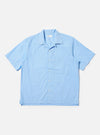 The Pale Blue Broadcloth Camp Shirt is shown laid flat against a plain white background. The short-sleeve shirt features a wide camp collar, a button-down front with a loop closure at the top, and a single large patch pocket on the left chest. The garment has a straight hem with side splits and is made from a smooth, light blue cotton fabric.