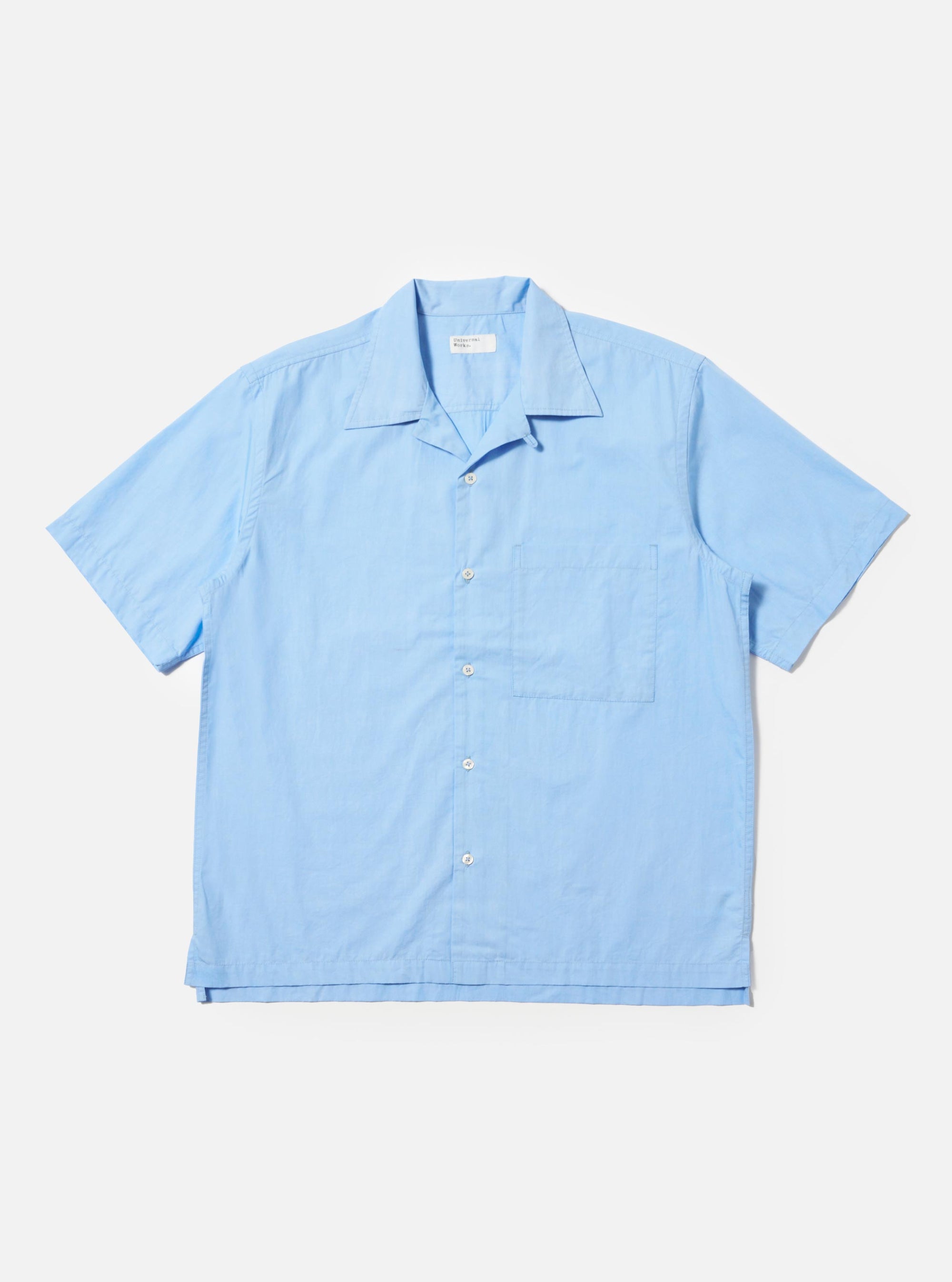 The Pale Blue Broadcloth Camp Shirt is shown laid flat against a plain white background. The short-sleeve shirt features a wide camp collar, a button-down front with a loop closure at the top, and a single large patch pocket on the left chest. The garment has a straight hem with side splits and is made from a smooth, light blue cotton fabric.