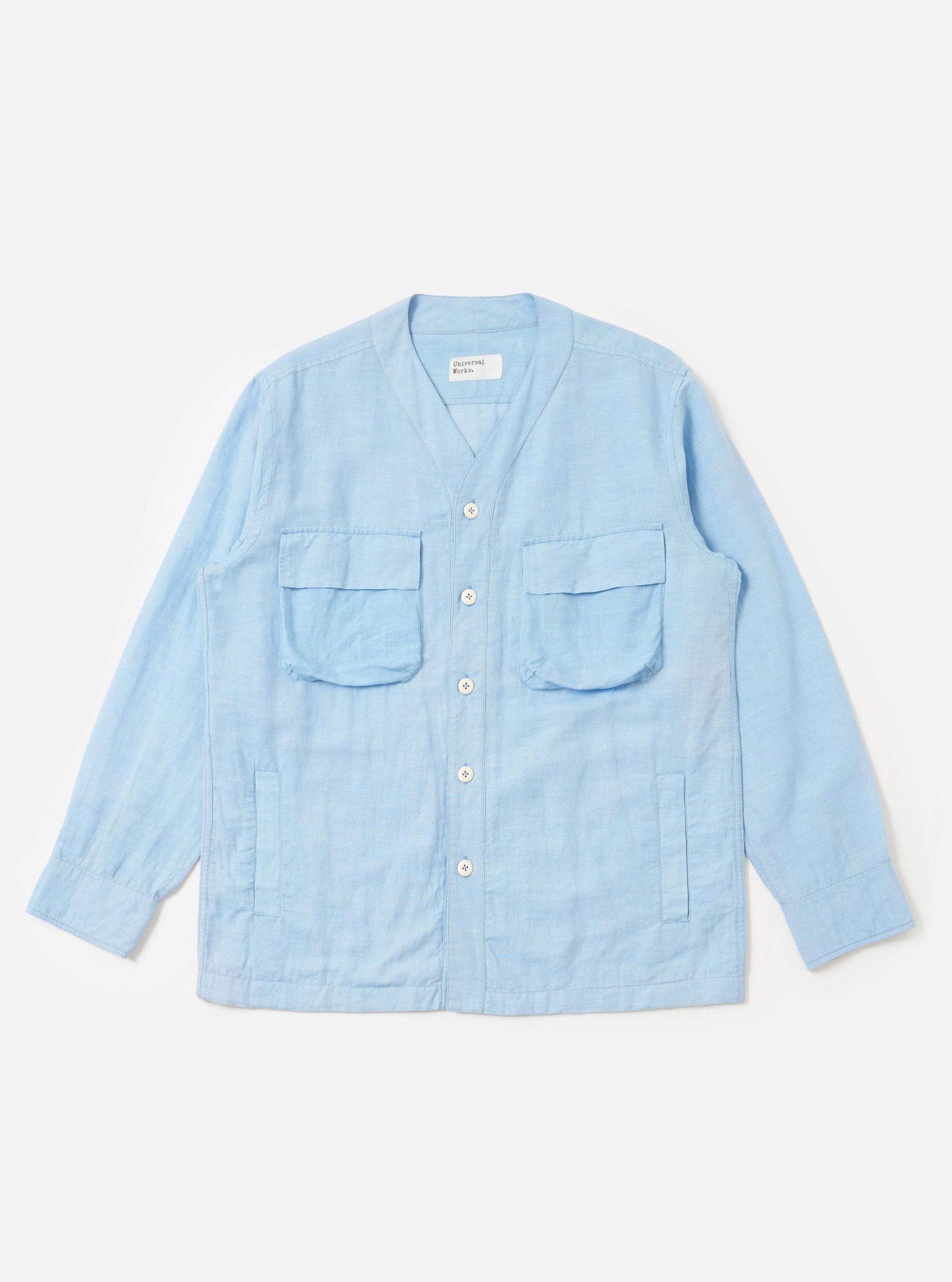 The Pale Blue Linen Cotton Shirting Parachute Shirt is displayed flat against a plain white background. This collarless, V-neck overshirt features a button-down front, two large 3D bellows pockets with buttoned flaps on the chest, and two vertical welt hand pockets near the waist. The garment is shown with long sleeves and a drawcord hem.