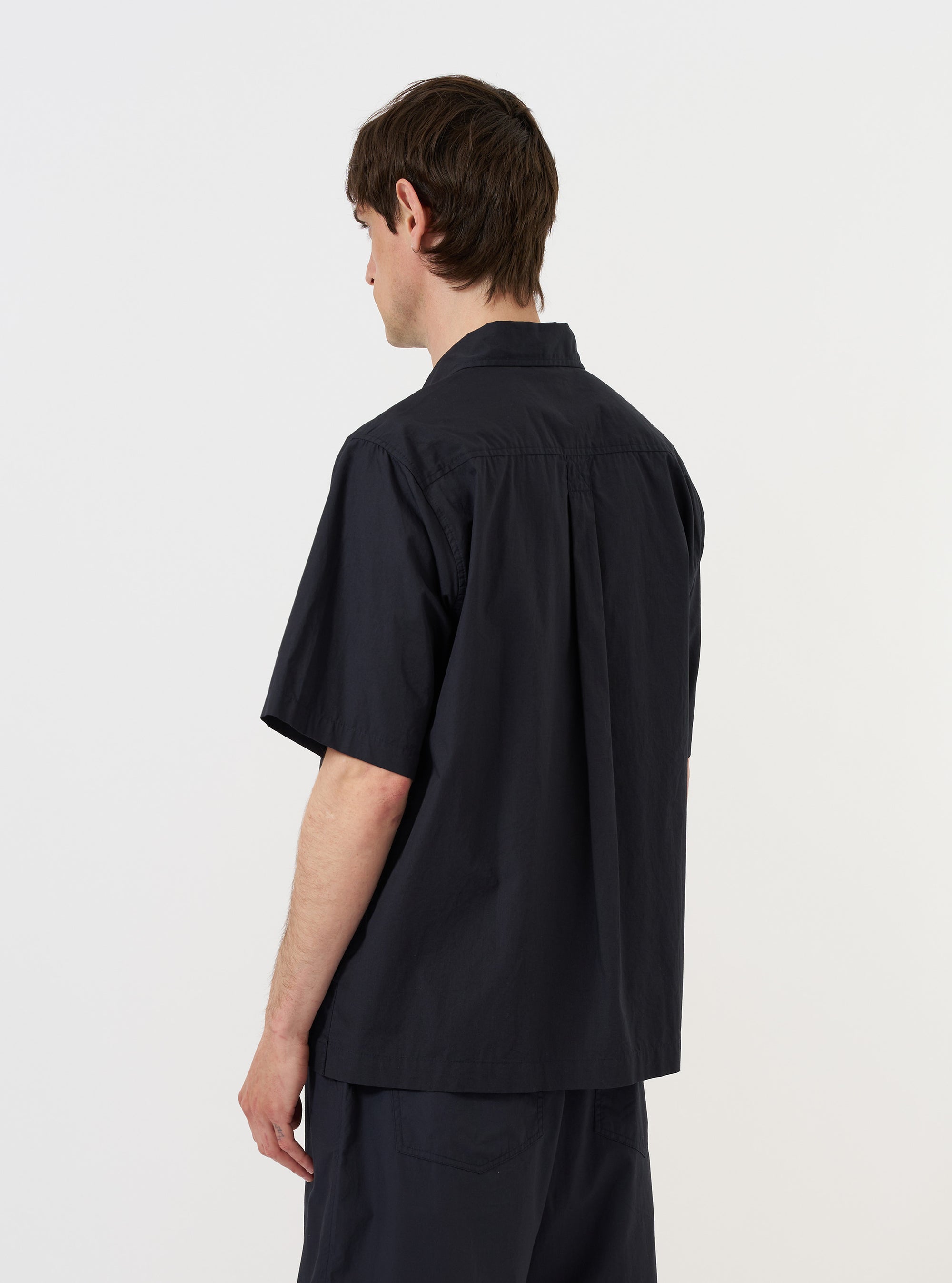 A model is wearing the Navy Broadcloth Ewan Shirt, shown from the back against a plain white background. The shirt is a navy blue pullover style with a zip detail at the neck, featuring a classic collar and a straight hem. The model is standing, and the image highlights the clean construction and smooth texture of the broadcloth fabric. No mannequin is present in the image.