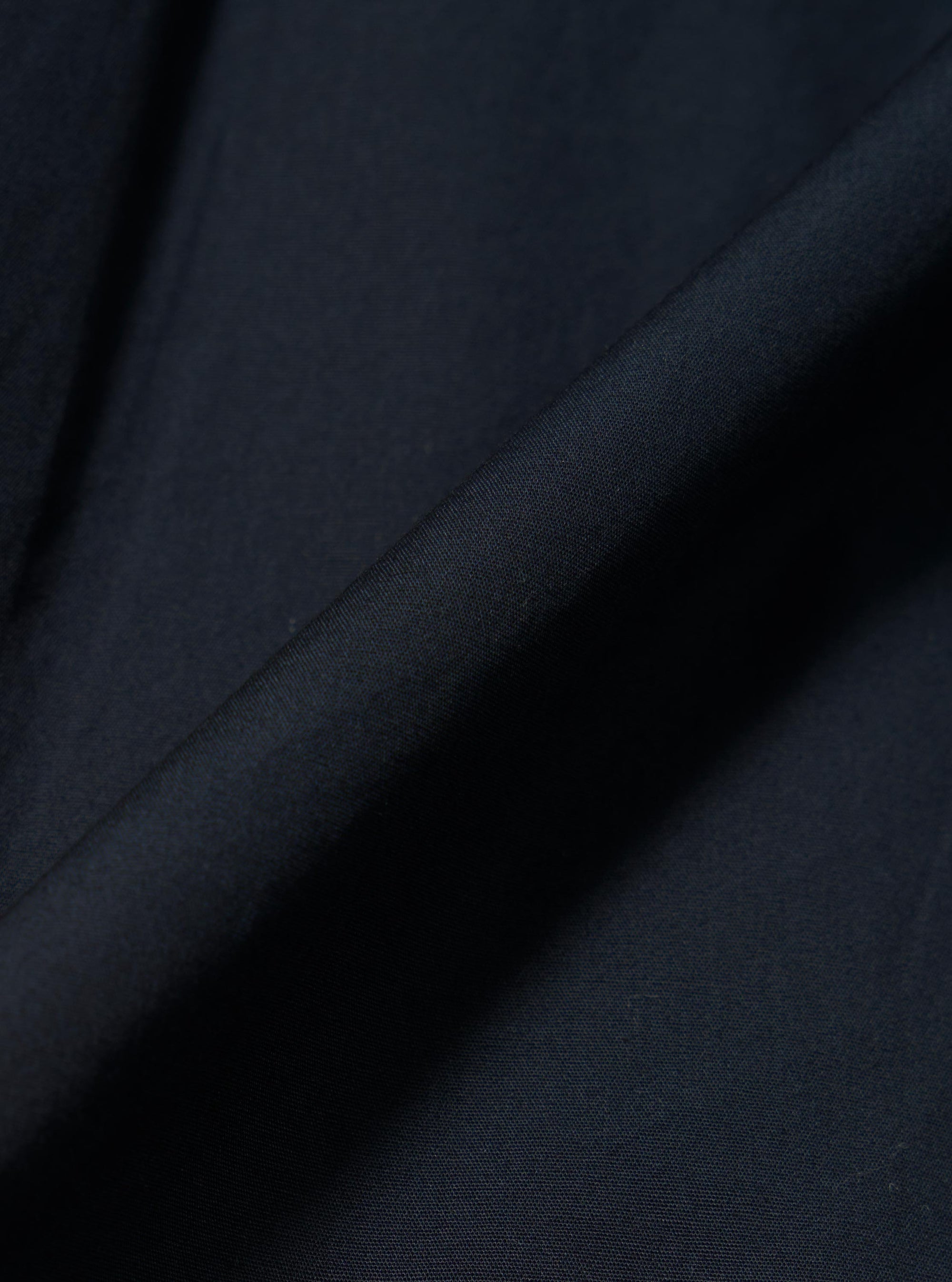 A close-up fabric swatch of the Navy Broadcloth Ewan Shirt highlights the smooth, tightly woven texture of the navy blue cotton material. The image focuses entirely on the fabric's fine grain and deep color.