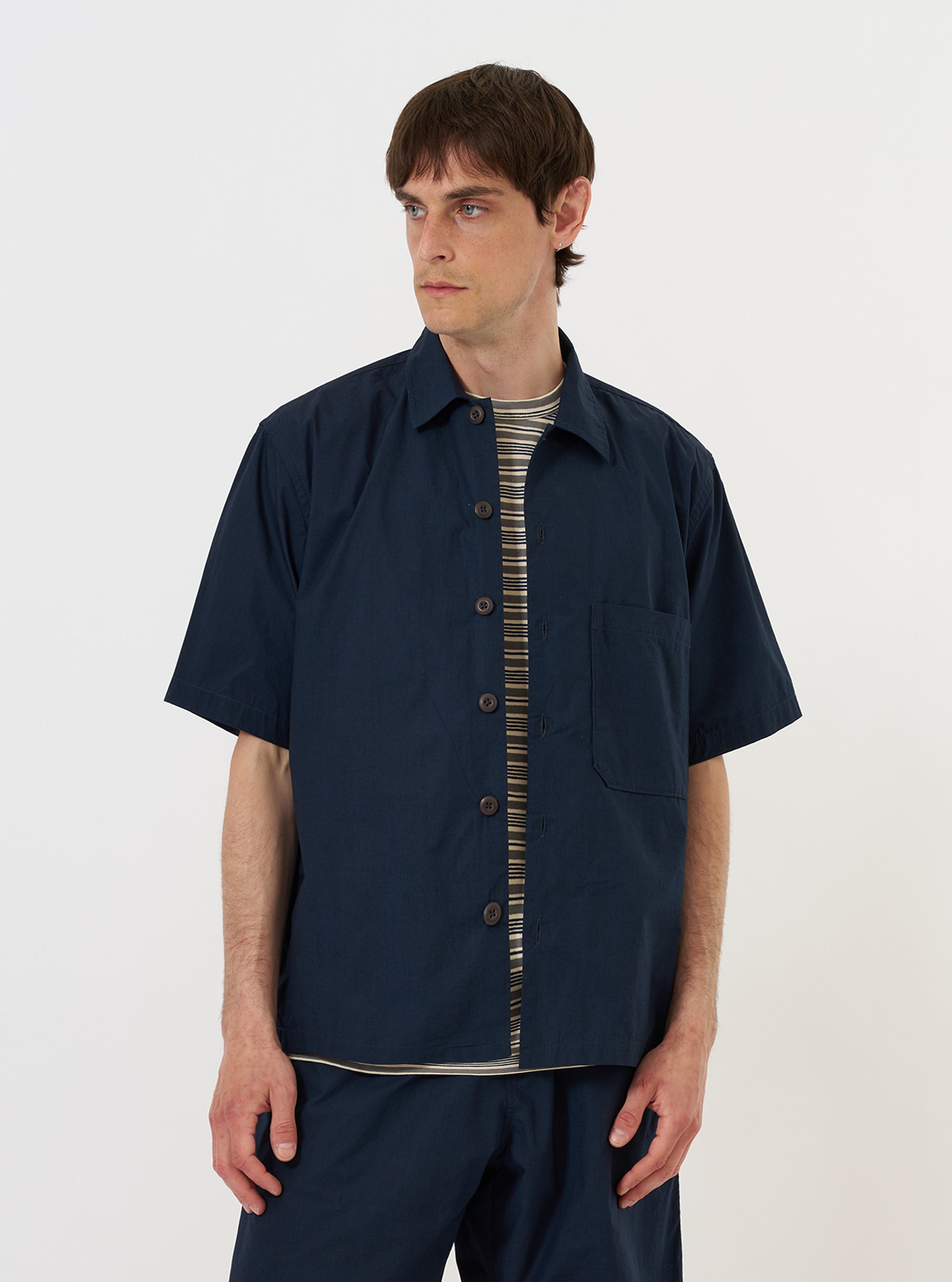 A model is wearing the Navy Paper Touch Cotton S/S Fatigue Shirt against a plain white background. The short-sleeved overshirt is shown from the chest up, featuring a classic pointed collar and two prominent chest pockets with buttoned flaps. The navy blue garment is worn over a white t-shirt, highlighting the crisp texture of the paper touch cotton.