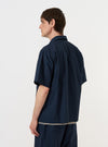 A model is wearing the Navy Paper Touch Cotton S/S Fatigue Shirt while standing against a plain white background. The image shows the short-sleeved overshirt from the back, highlighting the clean shoulder yoke and the crisp texture of the navy blue cotton fabric. The model is also wearing dark trousers. No mannequin is present in the image.