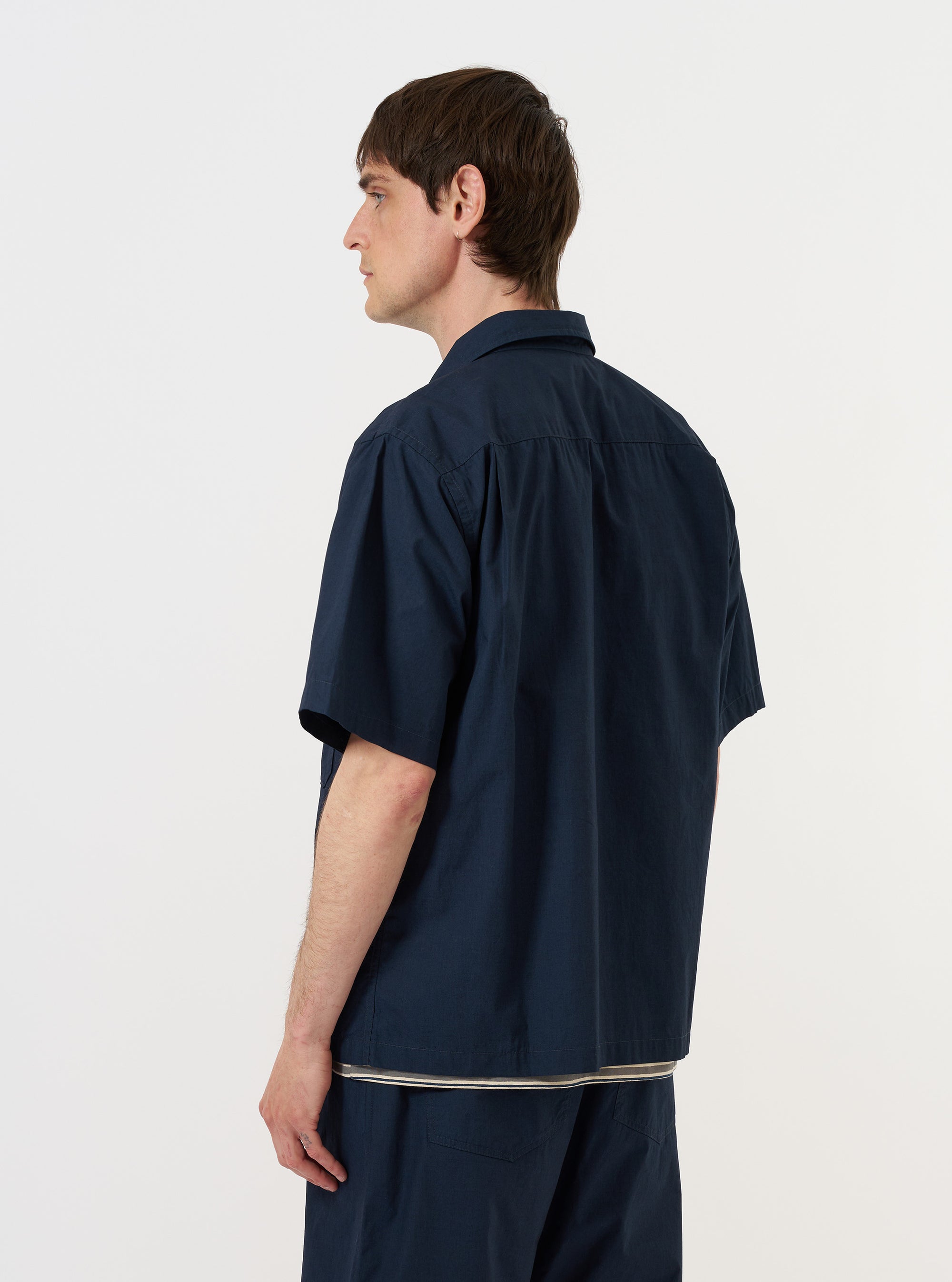 A model is wearing the Navy Paper Touch Cotton S/S Fatigue Shirt while standing against a plain white background. The image shows the short-sleeved overshirt from the back, highlighting the clean shoulder yoke and the crisp texture of the navy blue cotton fabric. The model is also wearing dark trousers. No mannequin is present in the image.