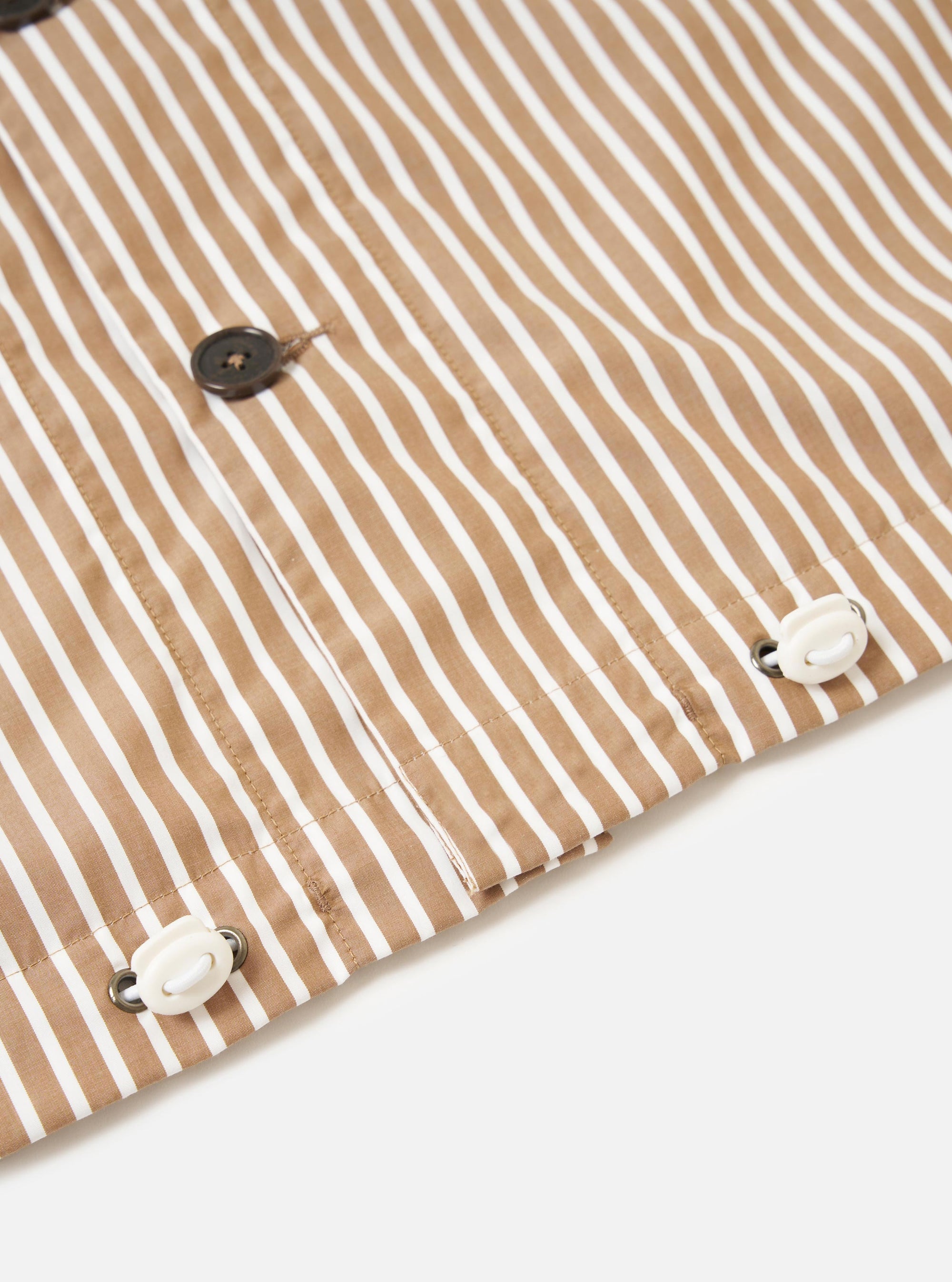 The Brown/White Cotton Stripe Z Tech Overshirt is shown from the back, laid flat against a plain white background. The garment features vertical brown and white stripes, a horizontal back yoke, and a central box pleat that adds volume to the silhouette. The hem is straight and incorporates an adjustable elasticated drawcord.