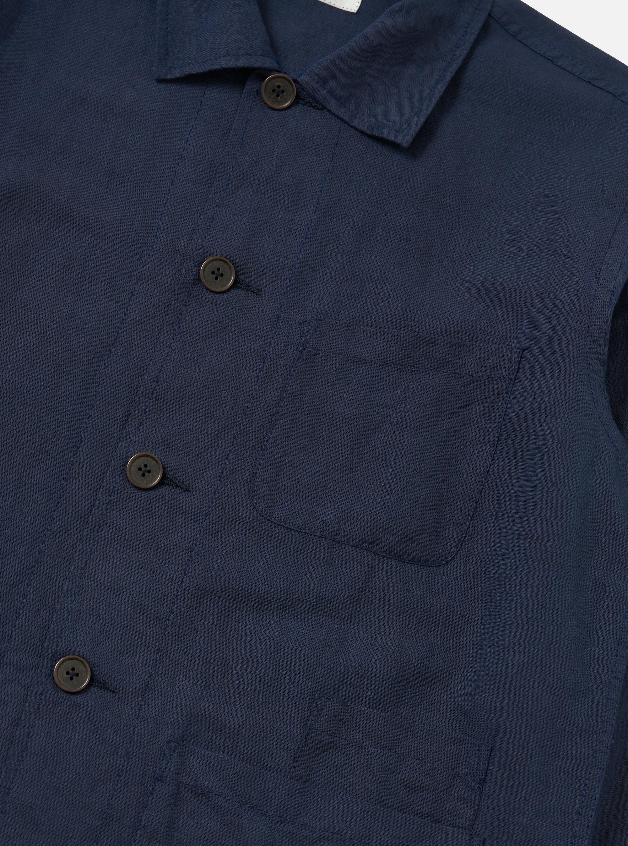 A model stands against a plain white background wearing the Navy Linen Cotton Shirting Bakers Overshirt over a white t-shirt. The navy overshirt features a classic collar, button-down front, and three large patch pockets on the front. The fabric has a visible linen texture, and the model has their hands by their sides.