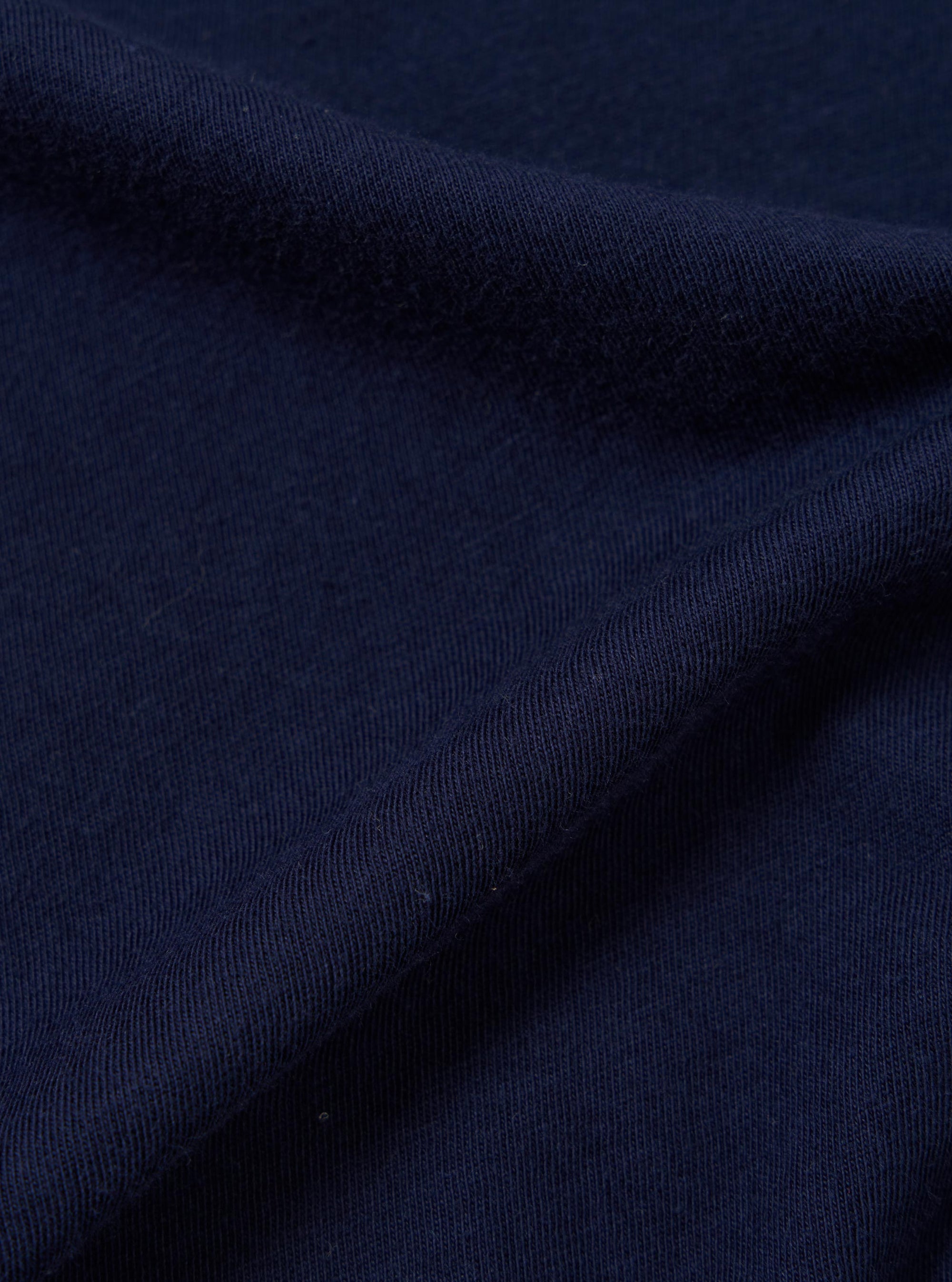 A close-up, square fabric swatch of the Navy Single Jersey Wish Wish You Were Here Tee is shown, detailing the textured navy blue cotton material. The image focuses on the fine knit of the single jersey fabric, showcasing its color and soft surface.
