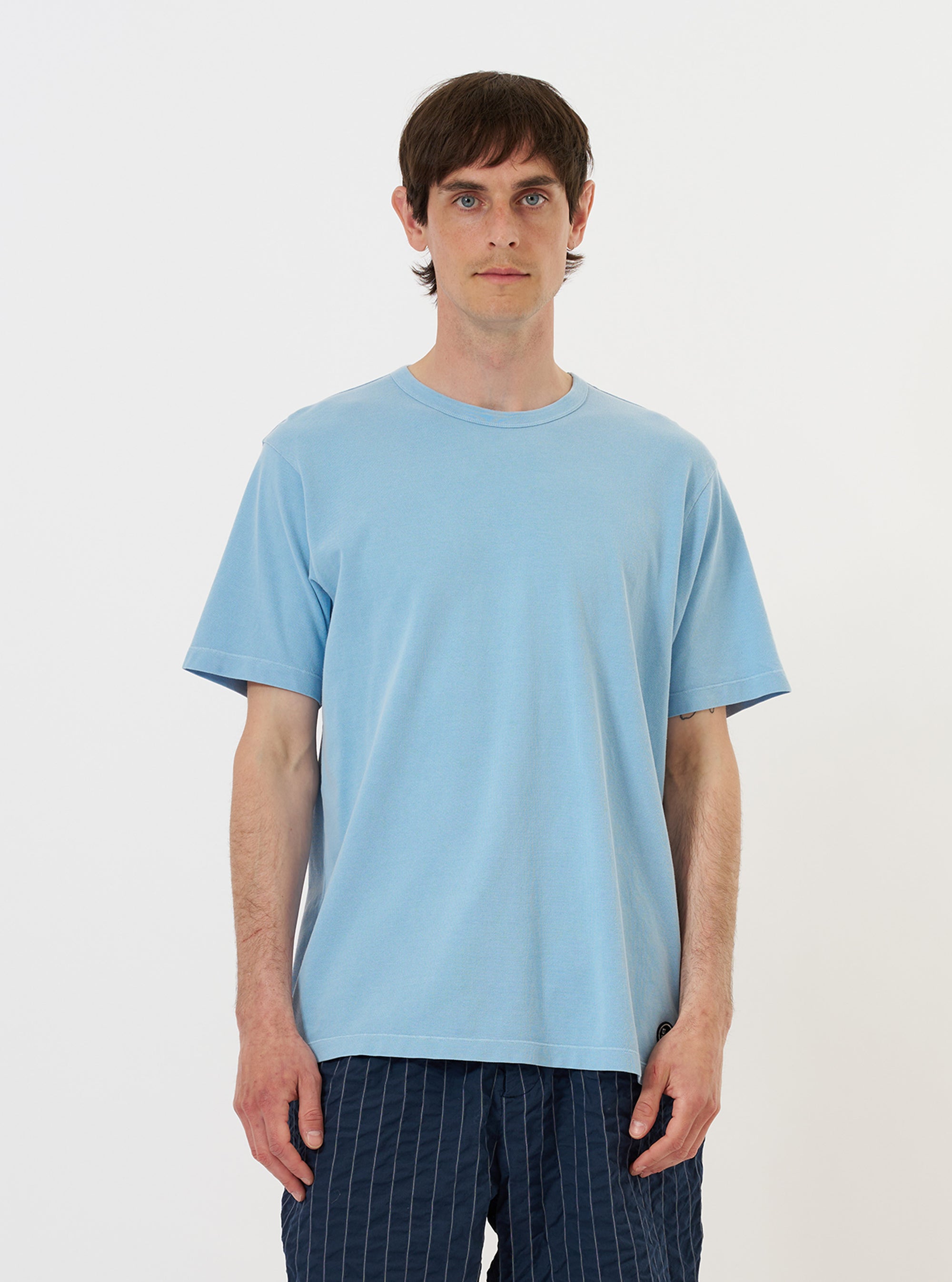 A model is wearing the Pale Blue Piquet Core Tee against a plain white background. The pale blue, short-sleeved t-shirt features a crew neck with a self-bound collar and a regular fit. The model is shown from the waist up, standing with a neutral pose, highlighting the texture of the piquet cotton fabric. No mannequin is present in the image.