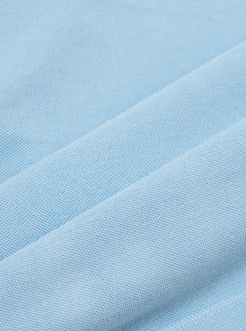 A close-up, square fabric swatch of the Pale Blue Piquet Core Tee highlights the detailed texture of the cotton piquet weave. The image captures the consistent, small-scale geometric pattern of the pale blue fabric.