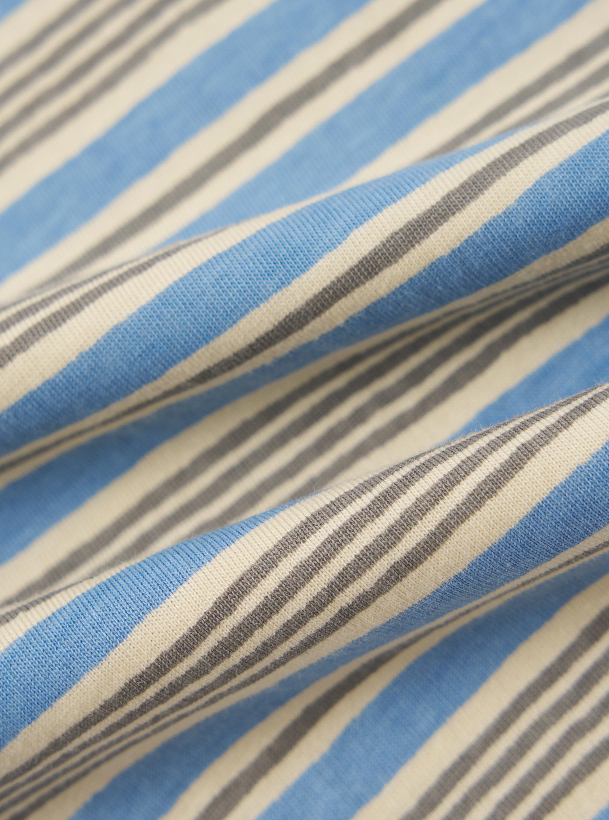 A close-up fabric swatch of the Pale Blue OS Stripe Jersey Core Tee displays a pattern of horizontal white and pale blue stripes. The image focuses on the fine, ribbed texture of the cotton jersey material.