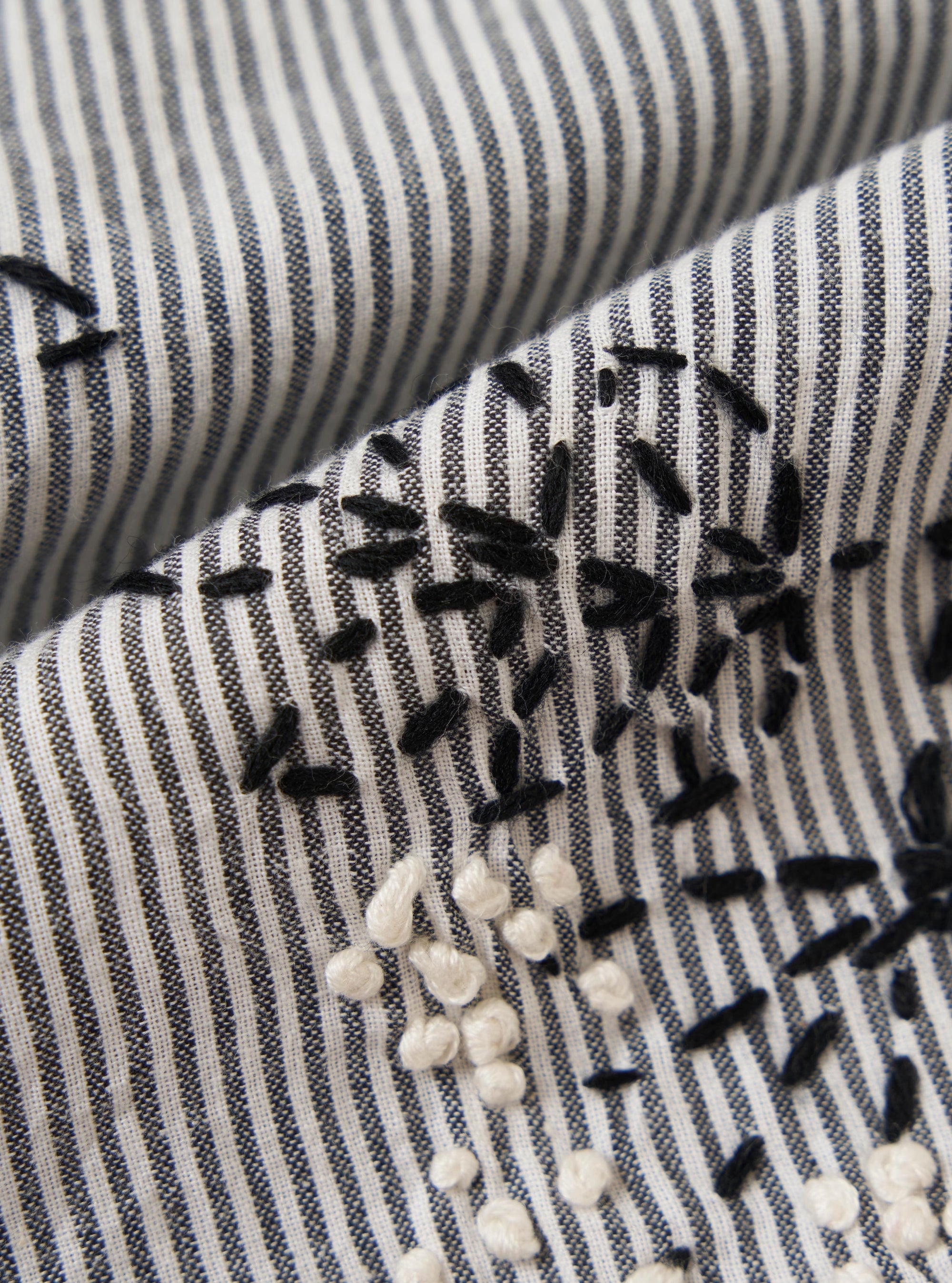 A square fabric swatch of the Grey Hand Embroidered Stripe Cotton Road Shirt shows a detailed close-up of the grey and white vertical striped fabric. Intricate hand-stitched floral embroidery in a darker thread is scattered across the material, highlighting the tactile texture of the cotton.