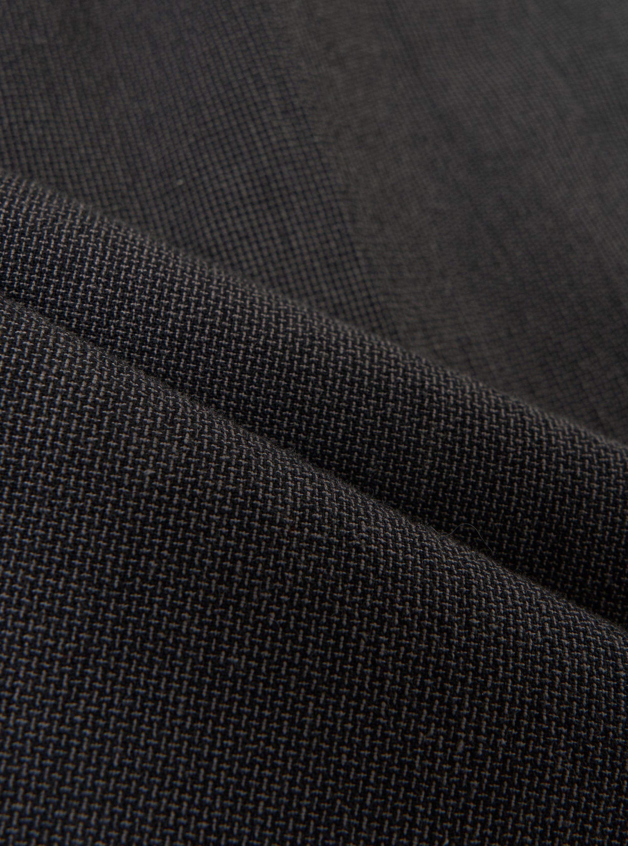 A detailed, close-up fabric swatch of the charcoal grey Seattle cotton material used for the Pleated Track Pant. The image highlights the fine basket weave and textured surface of the cotton fabric.