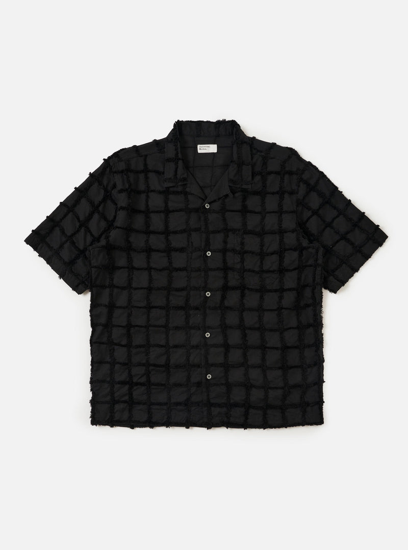 The Black 3D Check Road Shirt is displayed flat against a plain white background. The shirt features a short-sleeve silhouette, a relaxed camp collar, a button-down front, and a single chest pocket. The fabric has a textured 3D check pattern in black and grey.