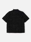 The Black 3D Check Road Shirt is shown from the back, laid flat against a solid white background. The image highlights the shirt's straight hem, short sleeves, and a horizontal yoke across the shoulders. The black fabric features a subtle, textured 3D check pattern throughout.
