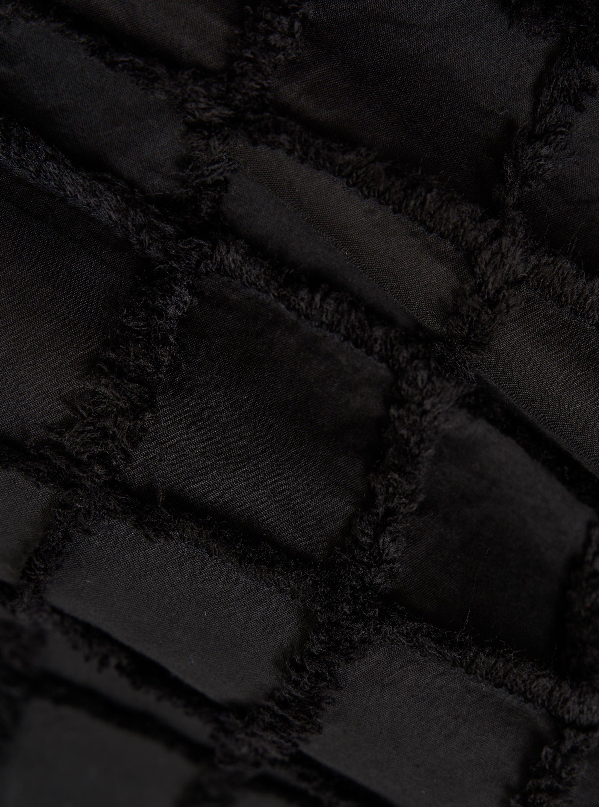 A detailed, close-up fabric swatch of the Black 3D Check Windcheater Jacket highlights a textured cotton material with a three-dimensional grid or checkered pattern in black and dark grey. The image focuses solely on the intricate weave and tonal contrast of the fabric.