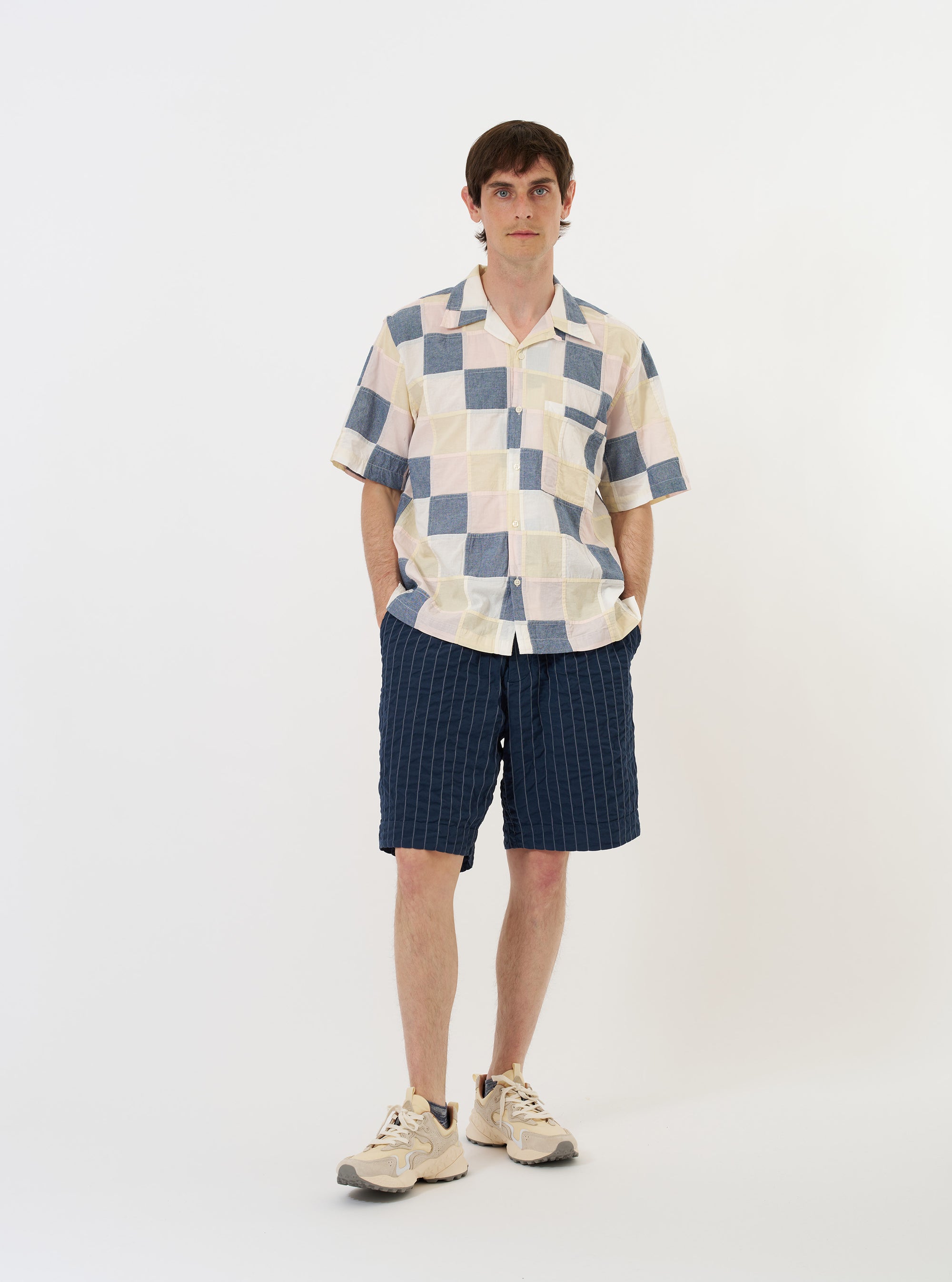 The Pastel Check Patchwork Camp Shirt is shown laid flat against a plain white background. The shirt features a short-sleeve silhouette with a multi-colored patchwork design consisting of various pastel check and plaid patterns in shades of blue, ecru, and pink. Key details include a relaxed camp collar, a full button-down front with a loop-close top button, and a large patch pocket on the left chest. The hem is straight with small side splits.