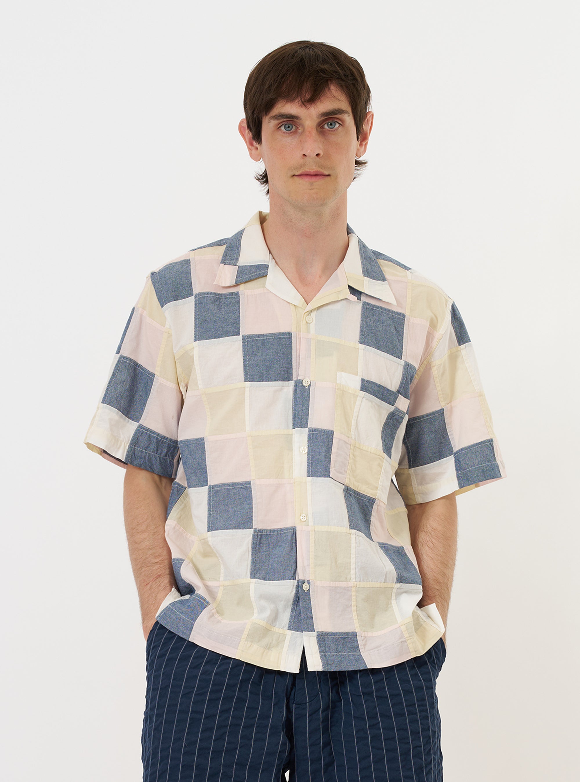 A model is wearing the Pastel Check Patchwork Camp Shirt against a plain white background. The short-sleeved shirt features a multi-colored patchwork design with various plaid patterns in pastel shades. It is styled with an open camp collar and a button-down front, featuring a single patch pocket on the chest. The model is shown from the waist up, highlighting the relaxed fit of the garment.