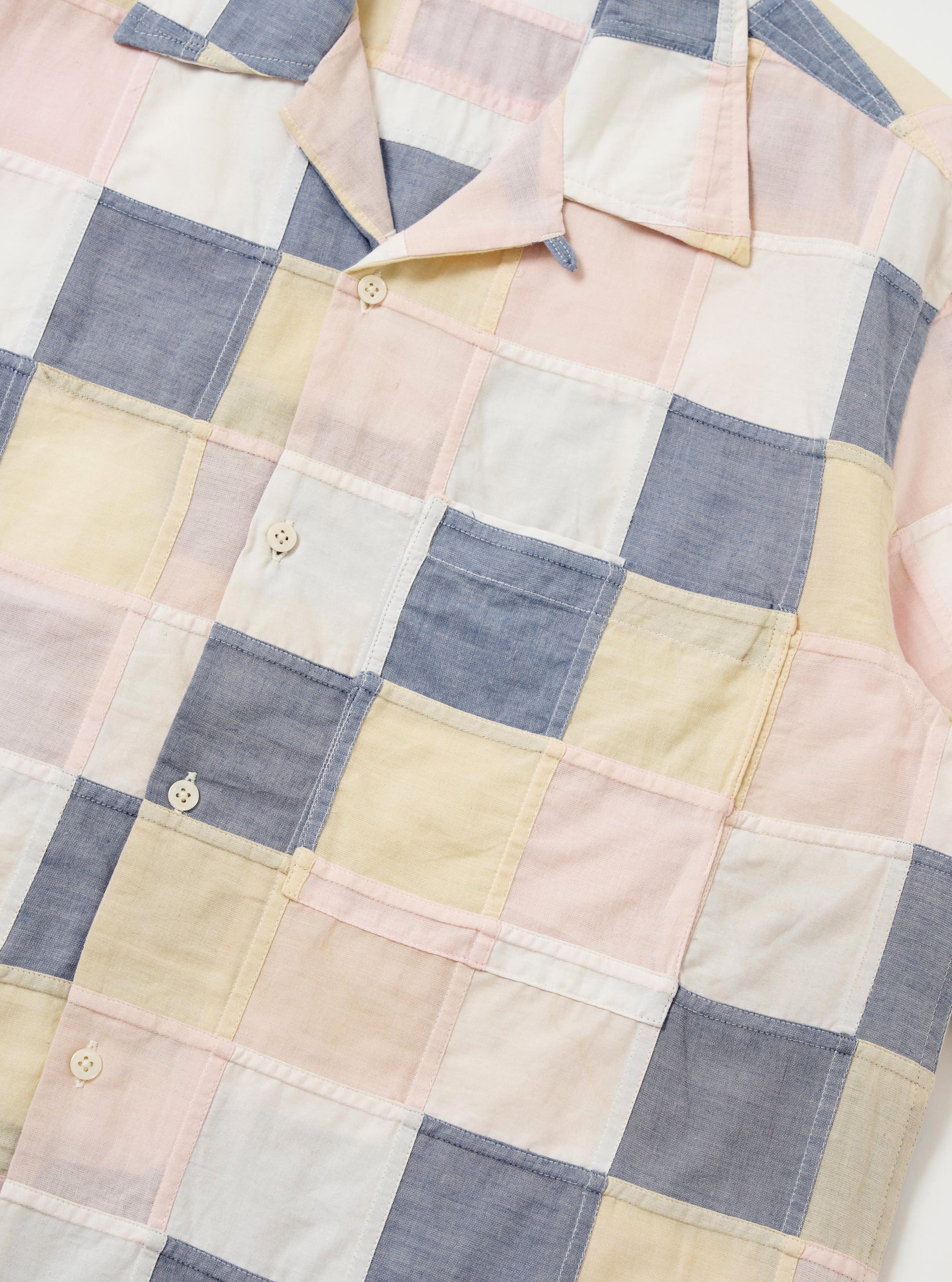 The Pastel Check Patchwork Camp Shirt is shown from the back, highlighting the construction of various multicolored plaid panels. The shirt features a horizontal back yoke with two small pleats and a straight hem with side splits. The garment is laid flat against a plain white background, and