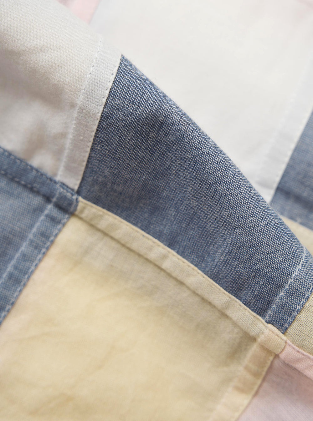 A close-up, square fabric swatch of the Pastel Check Patchwork Camp Shirt highlights a textured cotton material with a multi-colored patchwork check design. The pattern features various intersecting lines in pastel shades of blue, navy, and ecru.