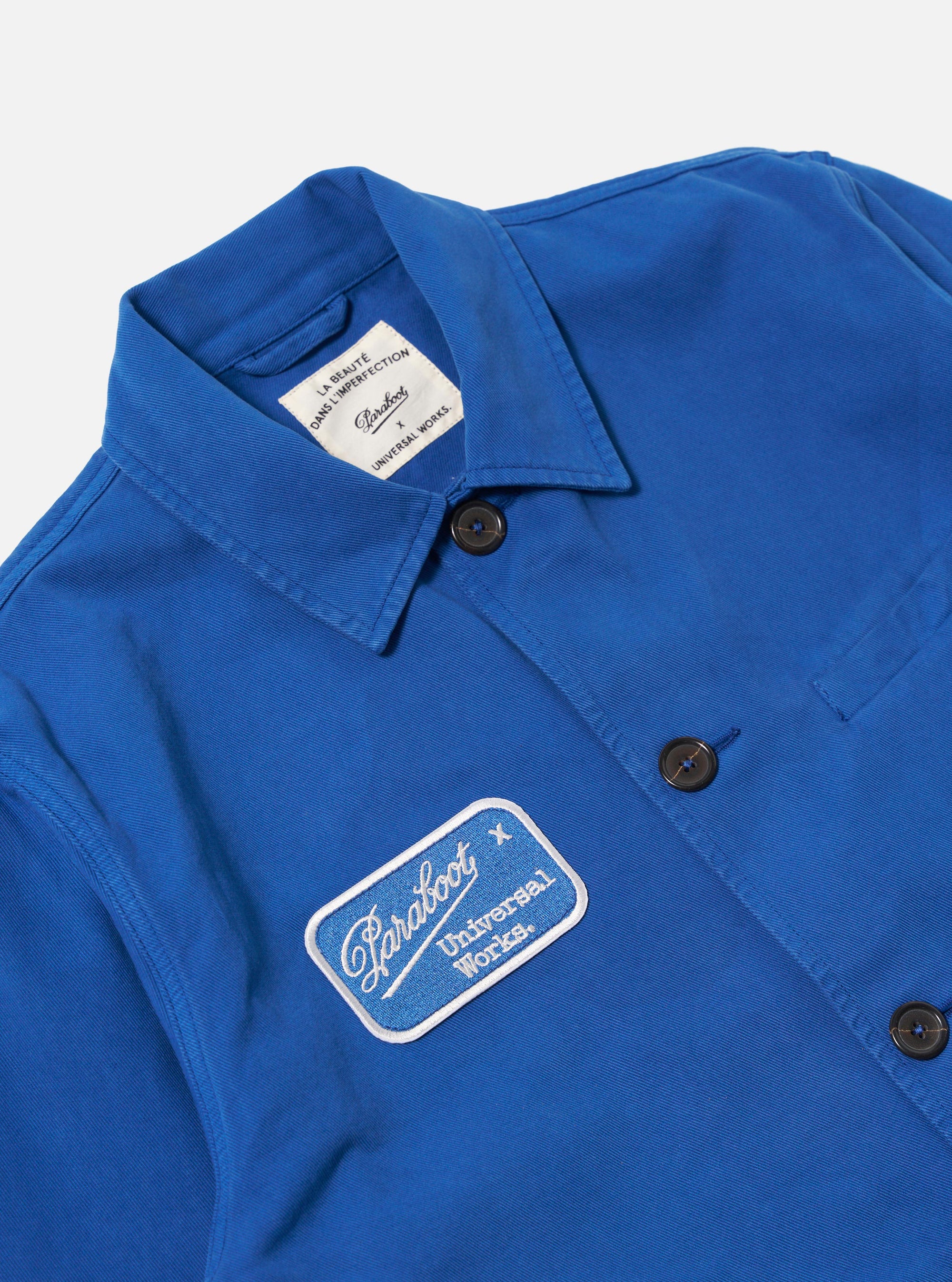 Close-up of the Paraboot x Universal Works Navy Derek Twill Warmus Jacket. Features a blue twill material, button closure, and branded patch. Shot on a flat surface.
