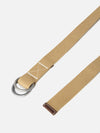 The Sand Canvas Webbing O Ring Belt is shown as a flat lay against a plain white background. The belt is made from a sand-colored woven canvas material and features two silver-tone O-ring buckles at one end. A dark brown leather trim is visible near the rings and at the tip of the belt.