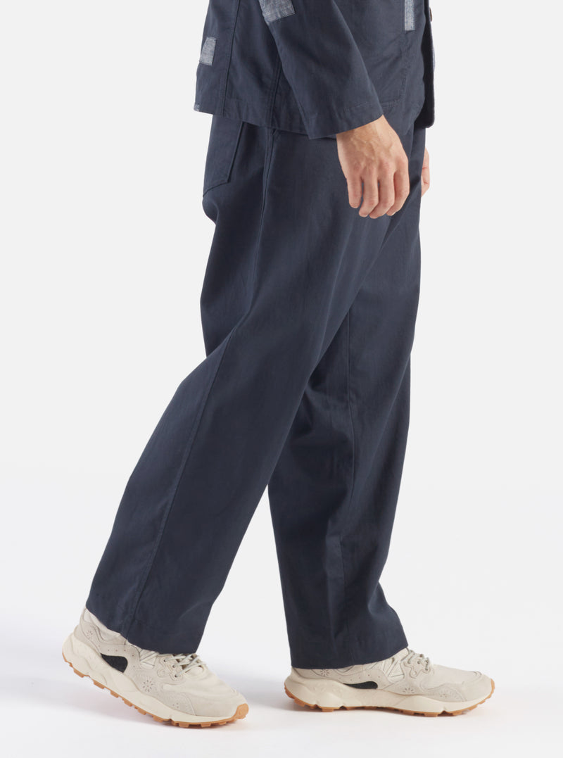 A model walks in a studio setting wearing the Navy Twill Duke Pant. The pants are navy and made of twill. The background is a bright white.