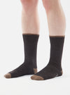 A close-up studio shot on a white background shows a person wearing Charcoal Alpaca Wool Sock. The sock is charcoal with a brown toe and cuff.
