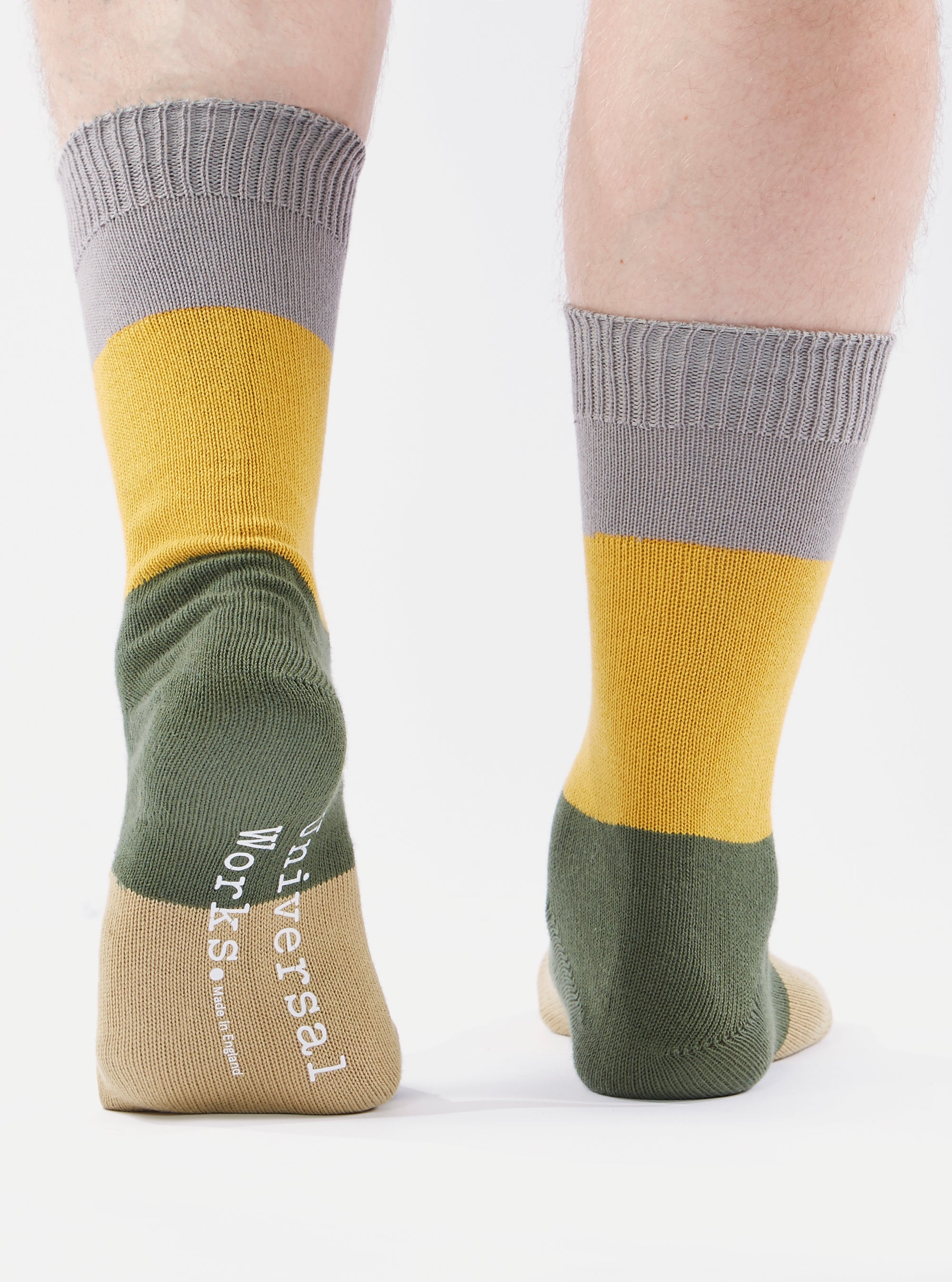 Close-up of the back of two legs wearing Grey/Gold Cotton Bold Stripe Sock on a white background. The socks have grey, gold, green and tan stripes.
