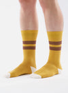 A model wears Yellow/Raisin Cotton Rib Sport Sock on a white background. The socks are yellow with raisin-colored stripes and white toes, made of ribbed cotton.