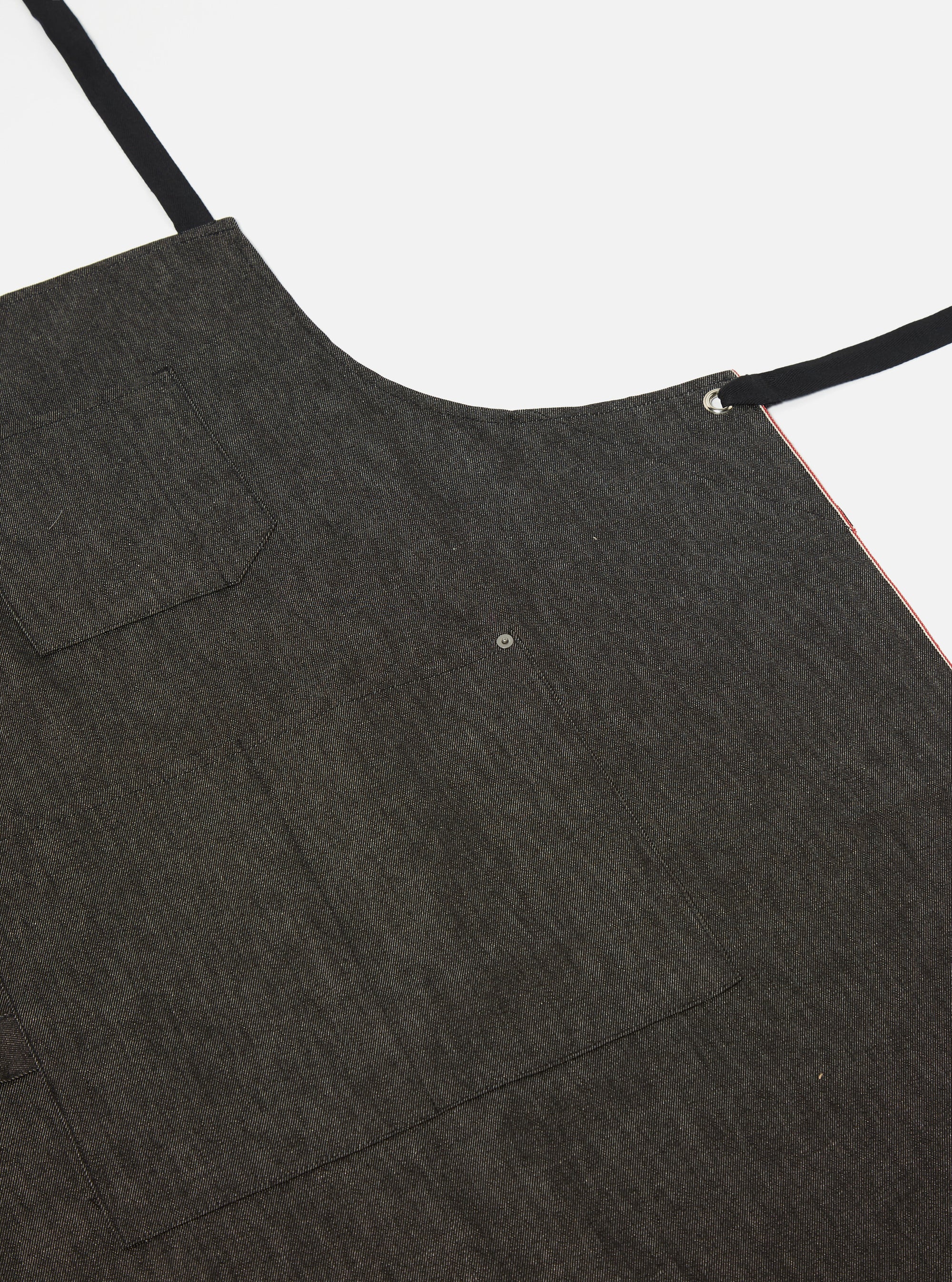 Close-up of a Black Selvedge Denim Apron on a white background. The apron has black straps and a small pocket. The selvedge edge is visible with a red line.