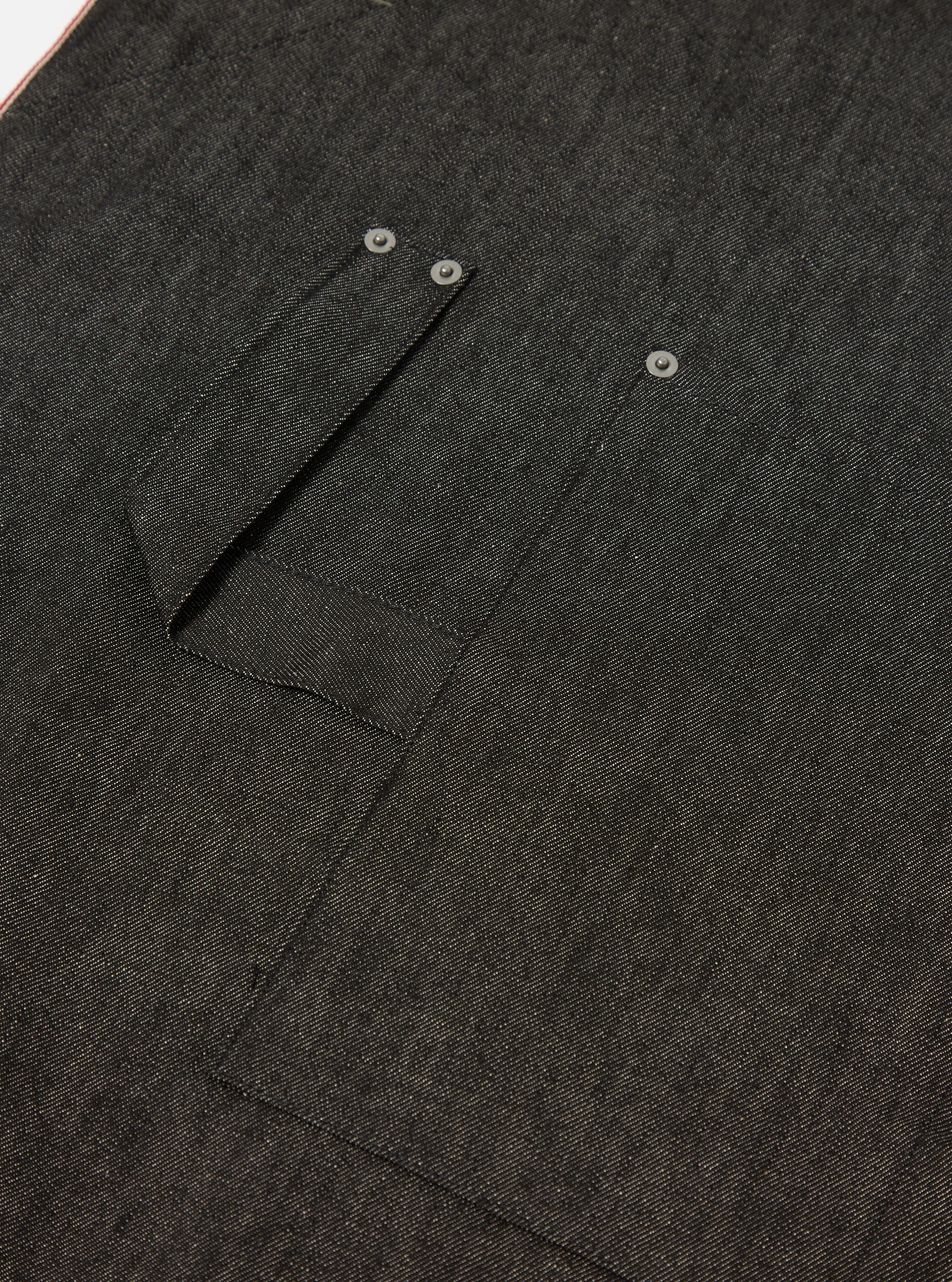 Close-up of the Black Selvedge Denim Apron showing the texture of the denim and pocket details. The apron is laid flat on a neutral surface.