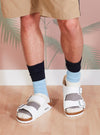 A person wearing the Navy/Sky Cotton Bold Stripe Sock with white sandals. The background is a pink wall with palm tree designs. The socks are cotton and fit snugly.