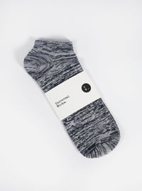 The Navy Slub Knit Ankle Slub Sock, packaged on a white surface. The sock is navy and white knit, with a paper label around it. 